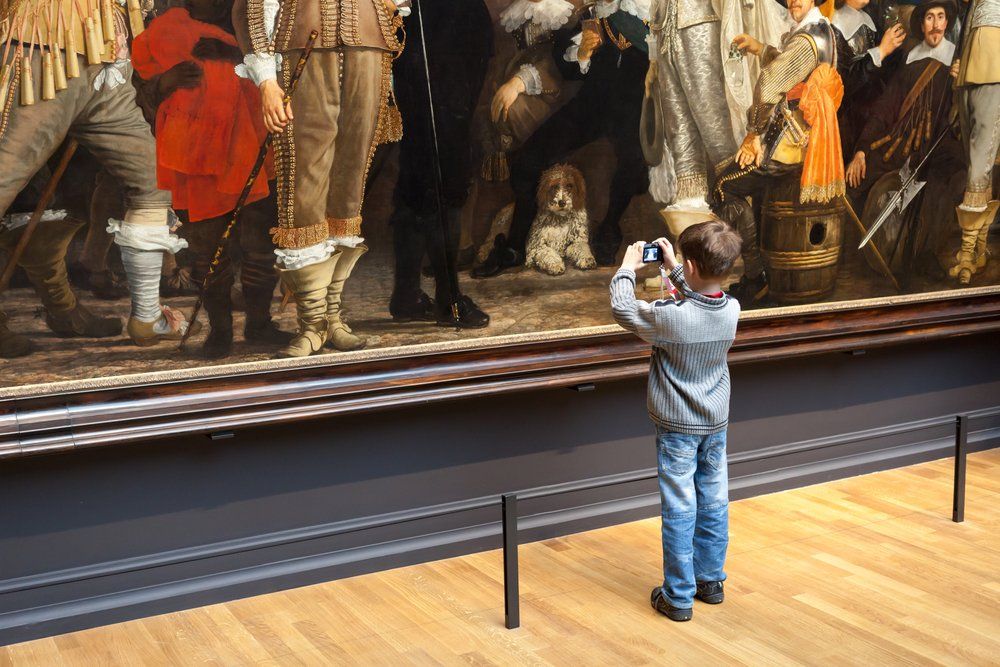 Niño fotografiando un gran cuadro de personas con ropas ornamentadas. Entorno museístico.