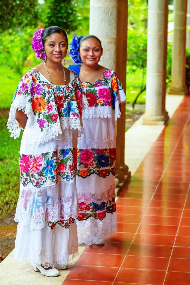 Two women in white embroidered dresses and flower hair accessories pose on a red-tiled patio.
