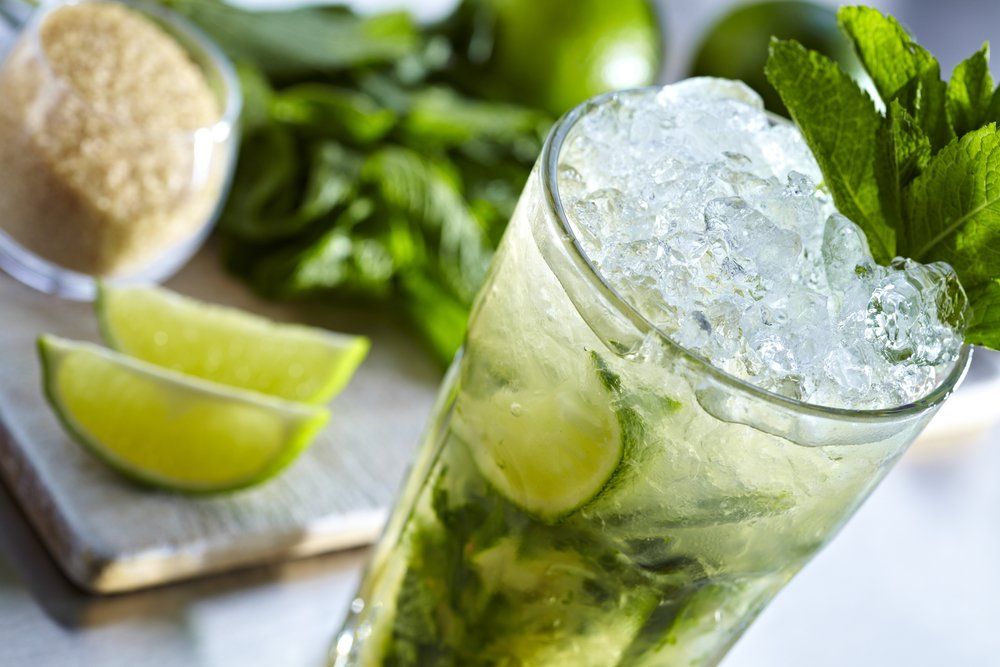 Cóctel mojito en vaso con hielo picado, gajos de lima, ramitas de menta y azúcar moreno sobre una superficie blanca.