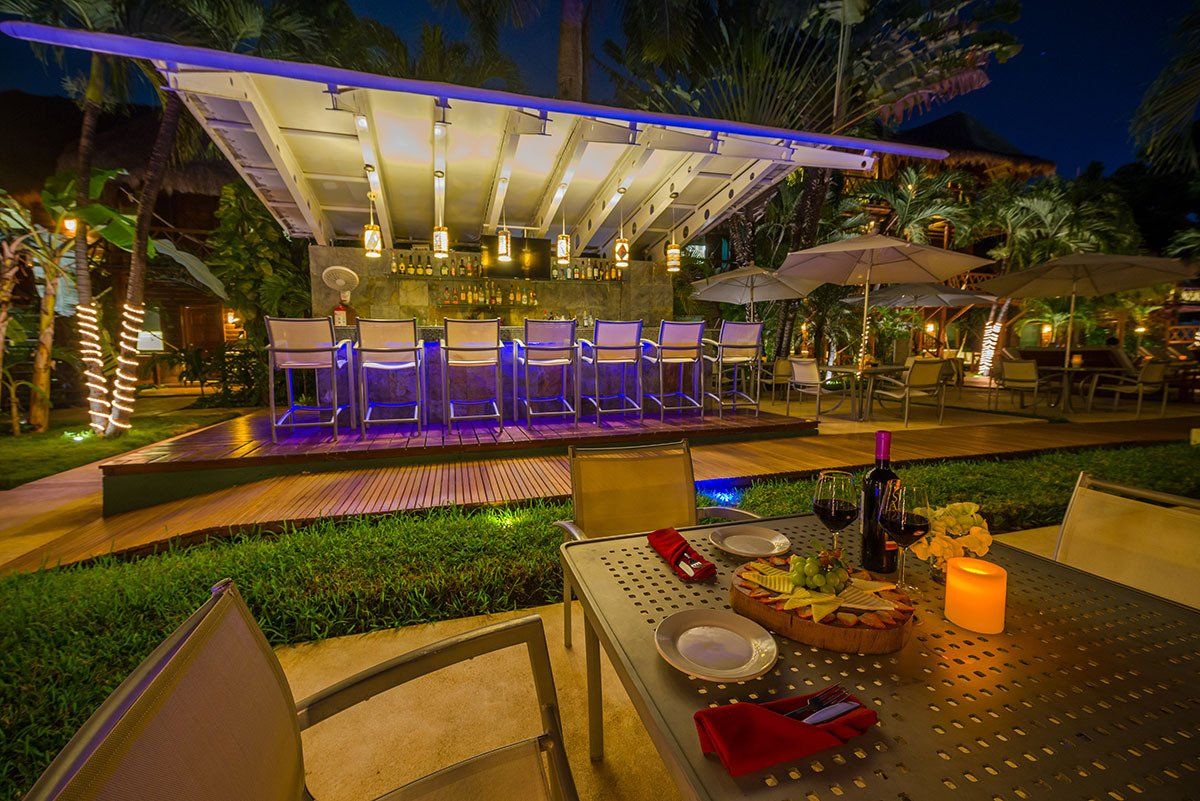 Outdoor bar at night with illuminated counter and seating, dining table set for two.