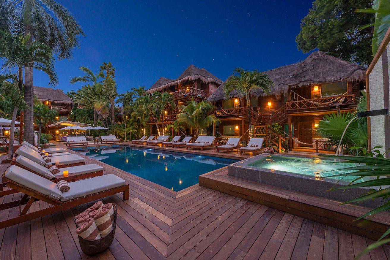 Poolside resort at night with a pool, hot tub, lounge chairs, and tropical buildings with thatched roofs.