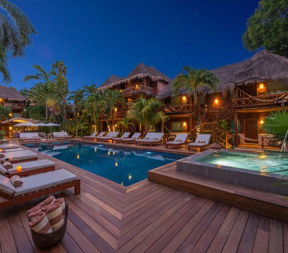 Poolside resort at night with lit buildings, wooden deck, palm trees, and lounge chairs.