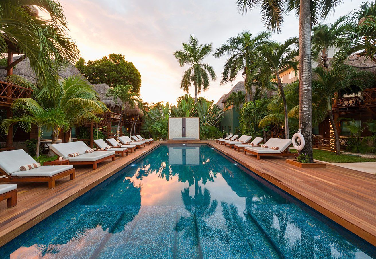 Swimming pool with lounge chairs, palm trees, and tropical resort buildings. Sunset sky reflected in water.