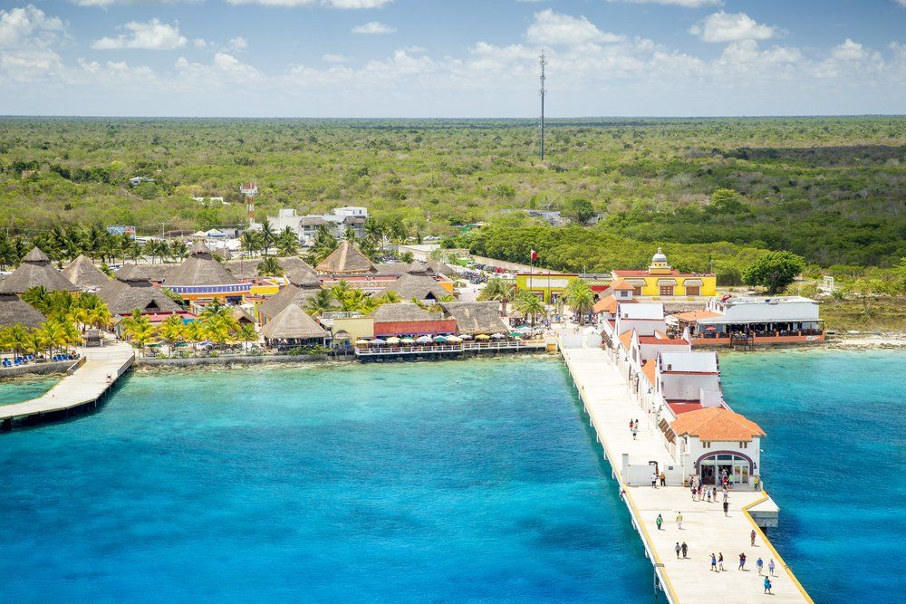 Pier extending into turquoise water, with buildings and greenery in the background, under a blue sky.