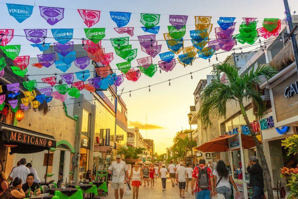 A pedestrian street in Playa del Carmen, Mexico, with colorful flags overhead, people walking, and shops.
