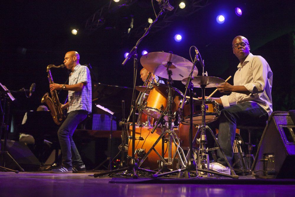 A jazz band performing on stage. Saxophonist playing, drummer at kit, lit by stage lights.