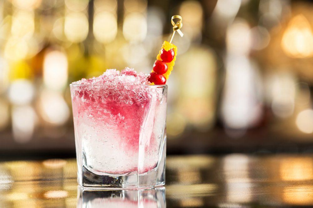 Cóctel en copa, con líquido rojo y hielo picado, adornado con una brocheta de fruta, sobre una barra borrosa.
