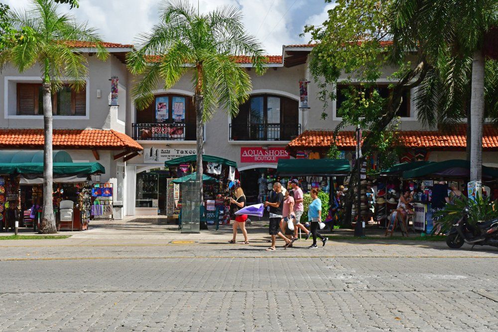 Shops with tiled roofs and palm trees line a sunny street with pedestrians walking.