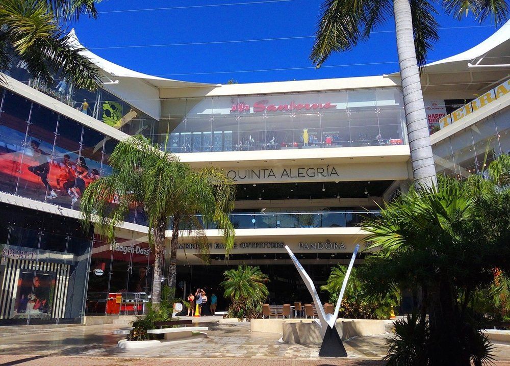 Mall entrance, Quinta Alegria. Modern architecture with glass and white accents, palm trees, and clear blue sky.