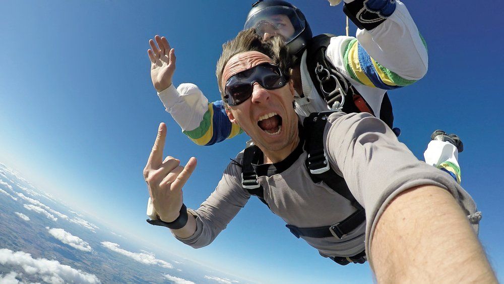 Man skydiving, giving a hand gesture, wide-eyed with open mouth, bright blue sky.