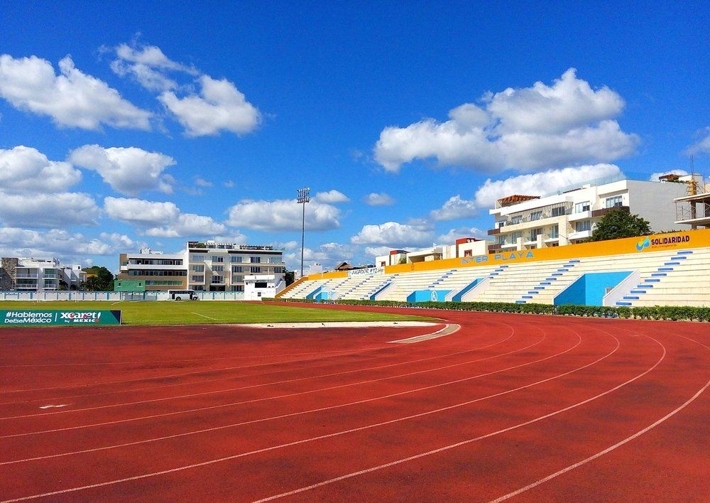 Red running track in a stadium with blue and yellow seating under a blue sky with clouds.