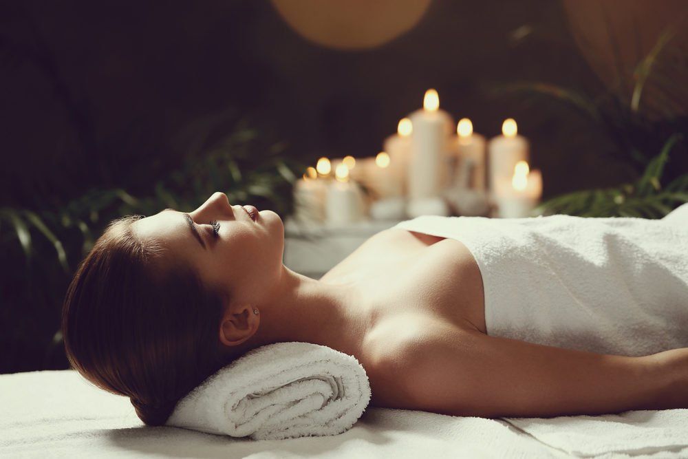 Woman lying on massage table, draped in white towel, in spa setting with candles.