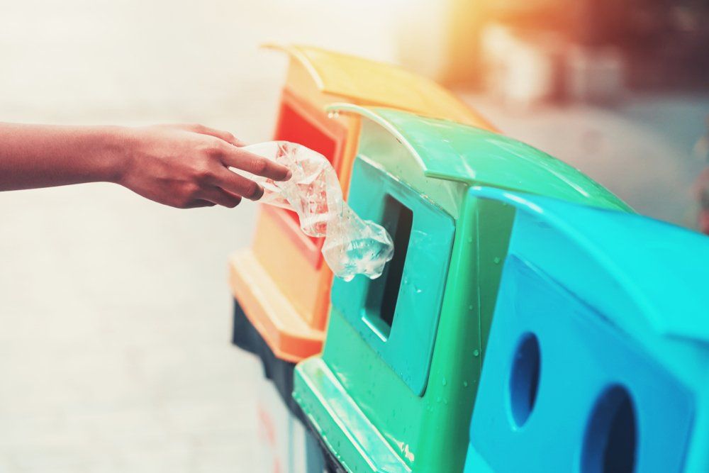Person placing a plastic bottle into a recycling bin. Colorful bins in a sunny outdoor setting.