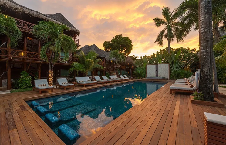 Piscina al atardecer, terraza de madera con tumbonas, palmeras y edificios tipo resort tropical.