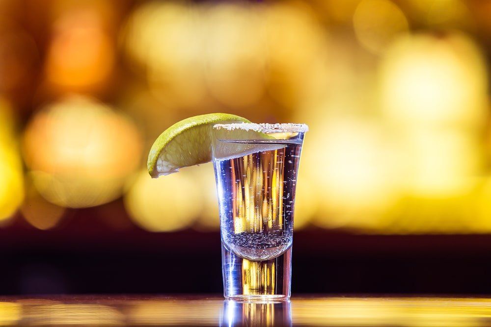 Shot de tequila con borde de sal y rodaja de lima, sobre un fondo dorado y borroso.