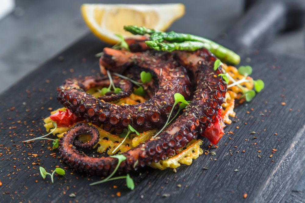 Pulpo a la parrilla con arroz amarillo, espárragos, limón y microvegetales sobre una tabla de madera negra.