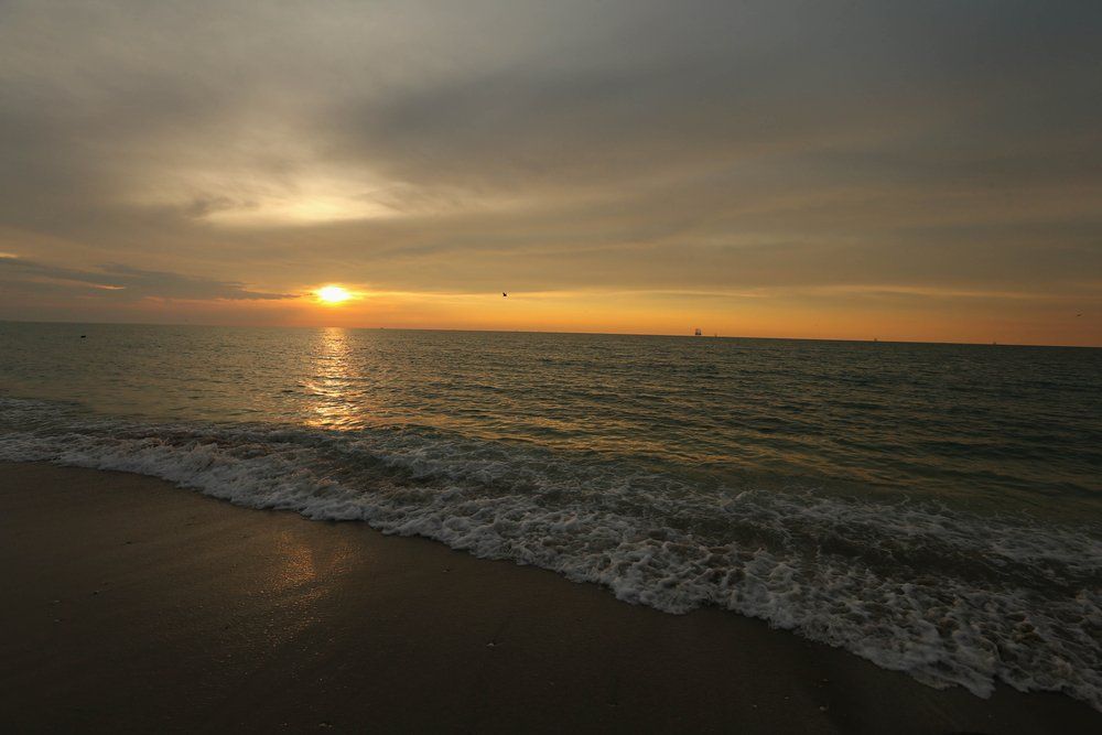 Sunset over ocean with waves gently lapping onto the sandy shore; orange and gold hues in the sky.
