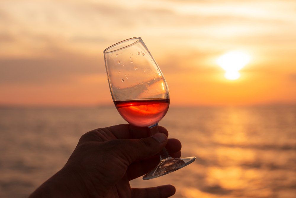Hand holding a glass of pink wine against a sunset over the ocean.