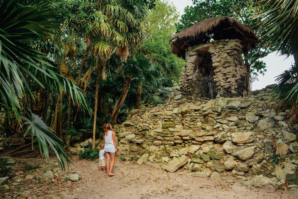 Woman and child walk towards a stone structure with thatched roof, surrounded by jungle foliage.