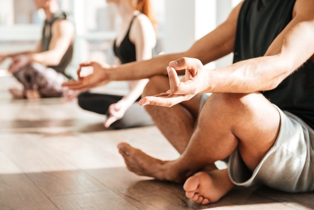 People meditating in a brightly lit room, hands in Gyan mudra, seated cross-legged on the floor.
