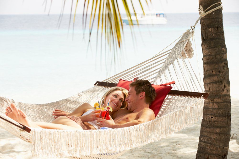 Pareja relajándose en una hamaca en una playa, tomando bebidas, con una palmera y un barco de fondo.