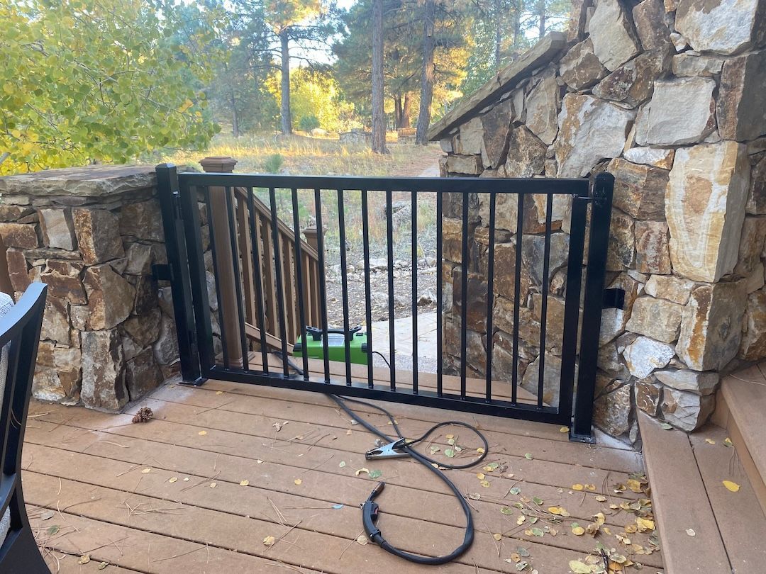 Black metal gate across a stone patio with a garden hose, surrounded by stone walls and trees outdoors