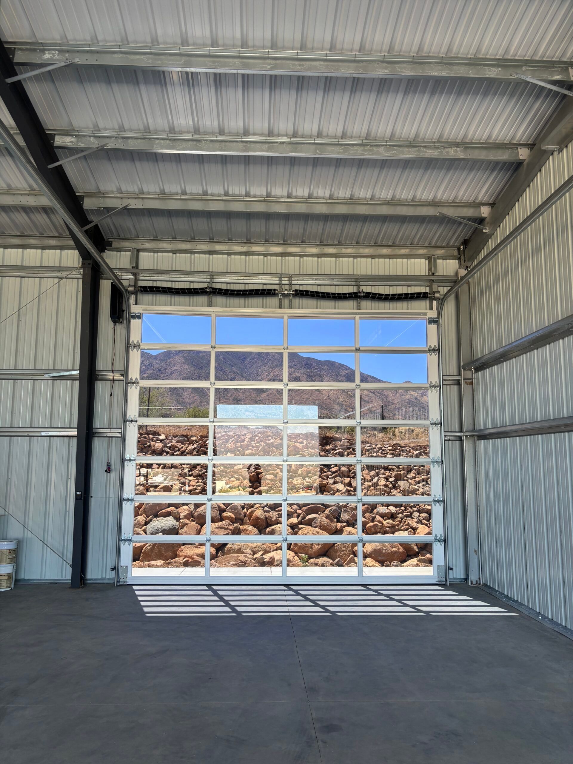 Empty metal garage with open door, showing a sunlit rock pile and blue sky outside.