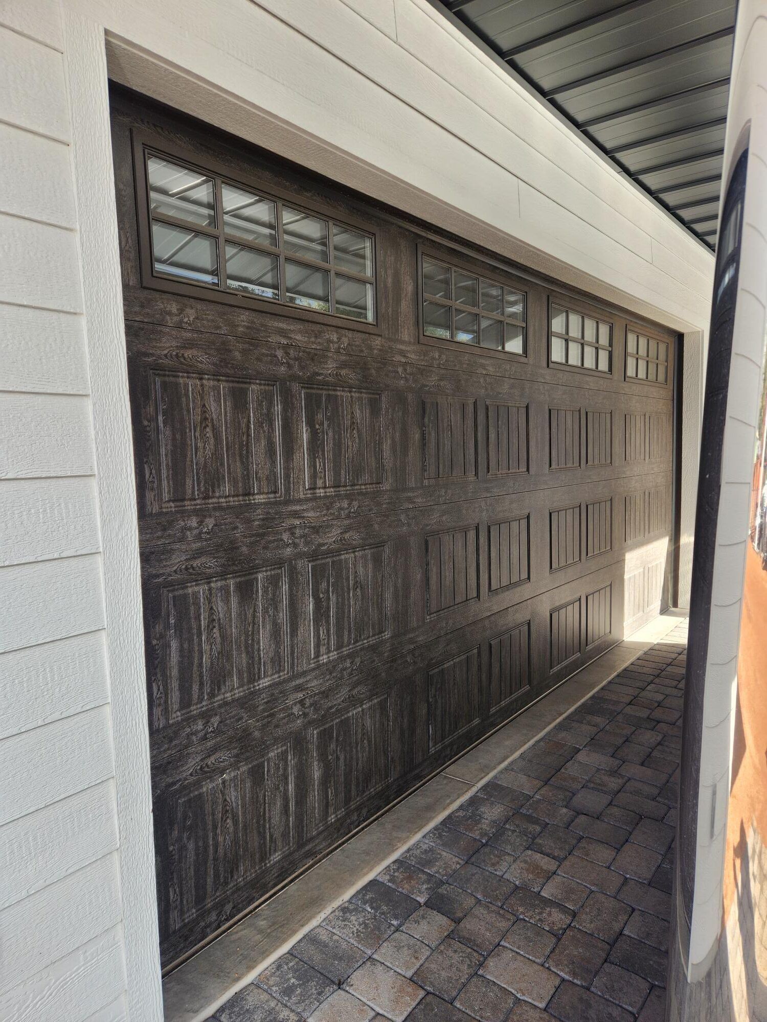Dark wood garage door with small windows, beside a white wall and brick driveway under a narrow overhang