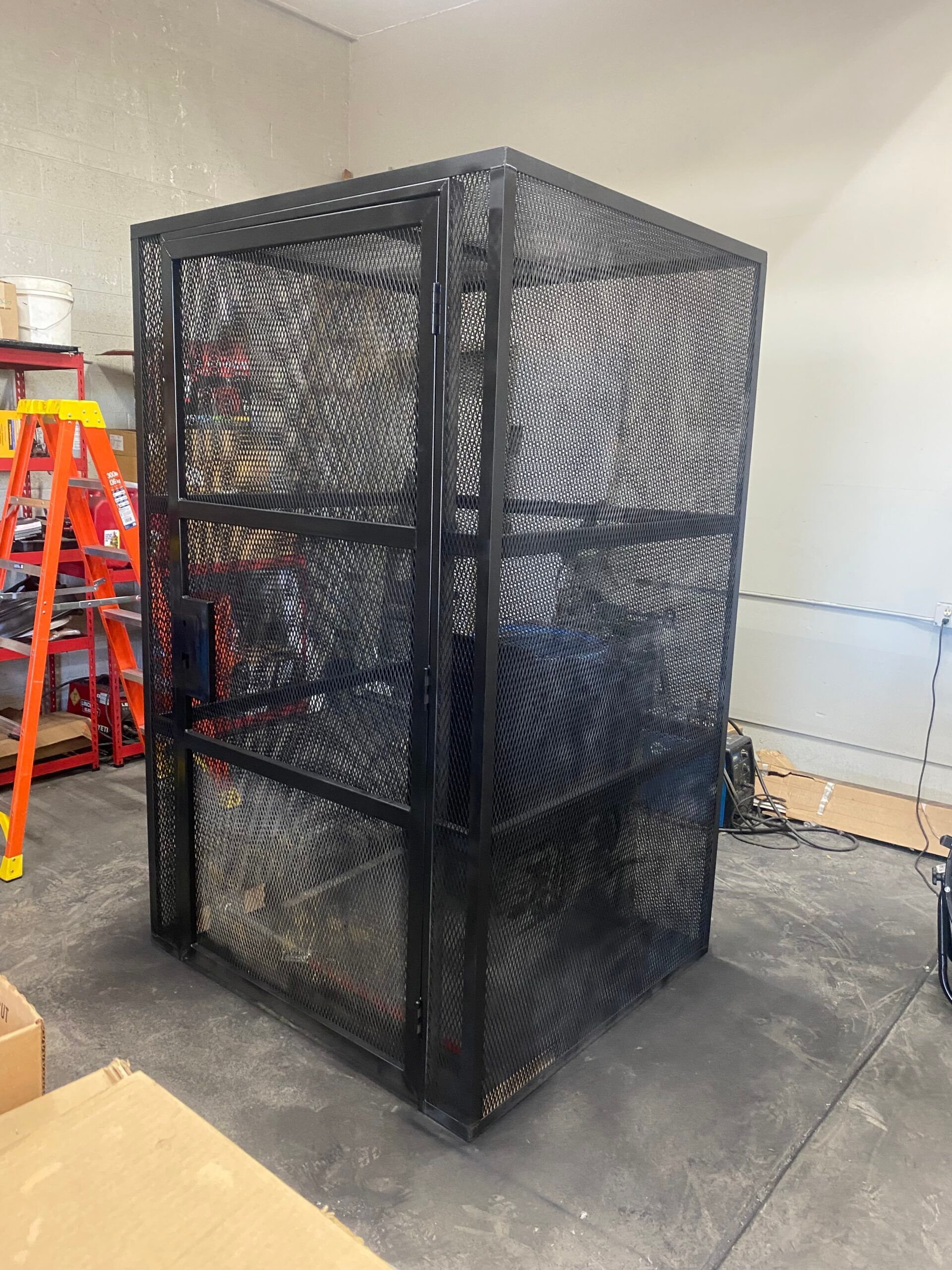Black metal mesh storage cage in a warehouse, standing on a concrete floor near a red ladder.