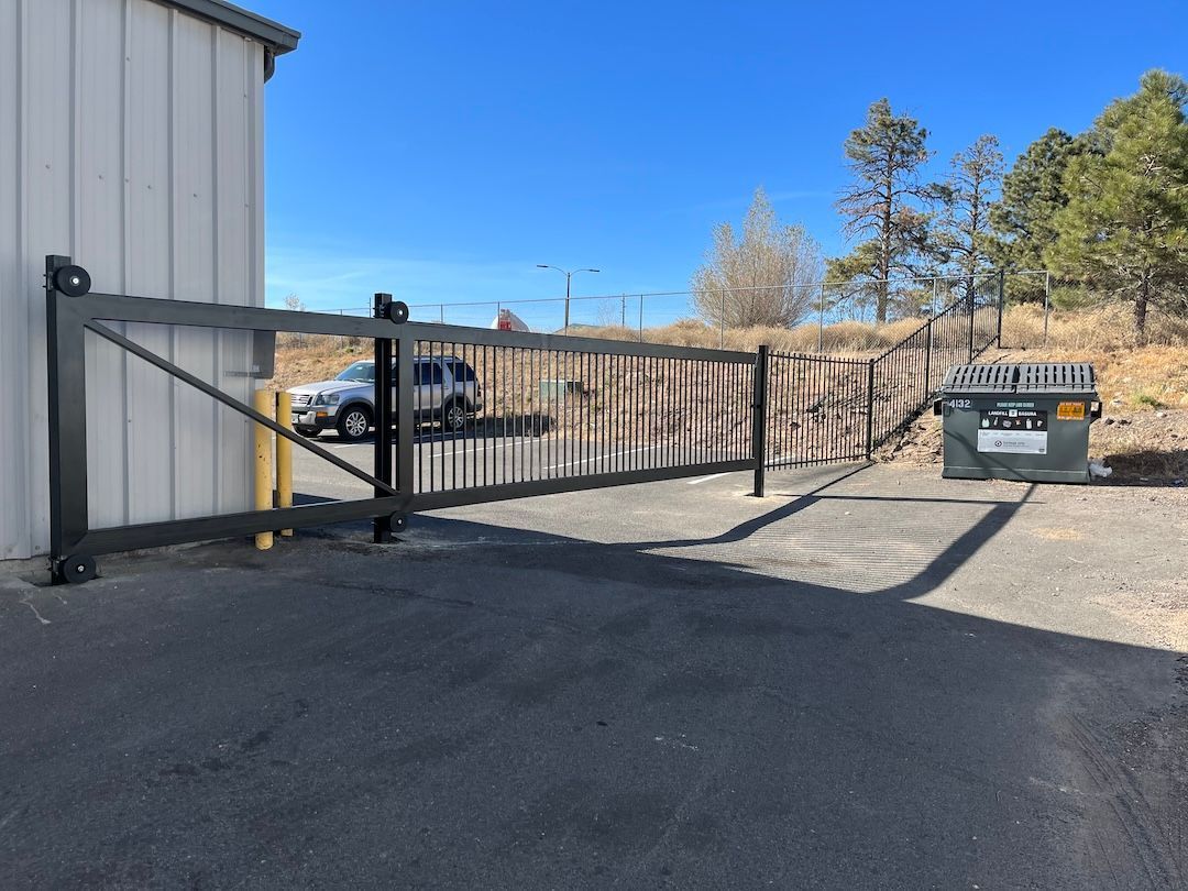 Closed metal gate across a driveway beside a white building, with trees and a dumpster in the background