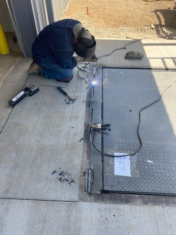 Welder crouched on concrete, welding a metal gate or grate beside a doorway, with sparks visible