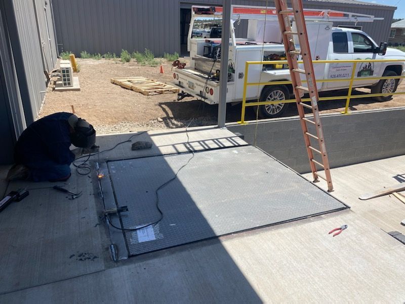 Worker kneels beside a large gray metal panel on a concrete pad, with a ladder and utility truck nearby.