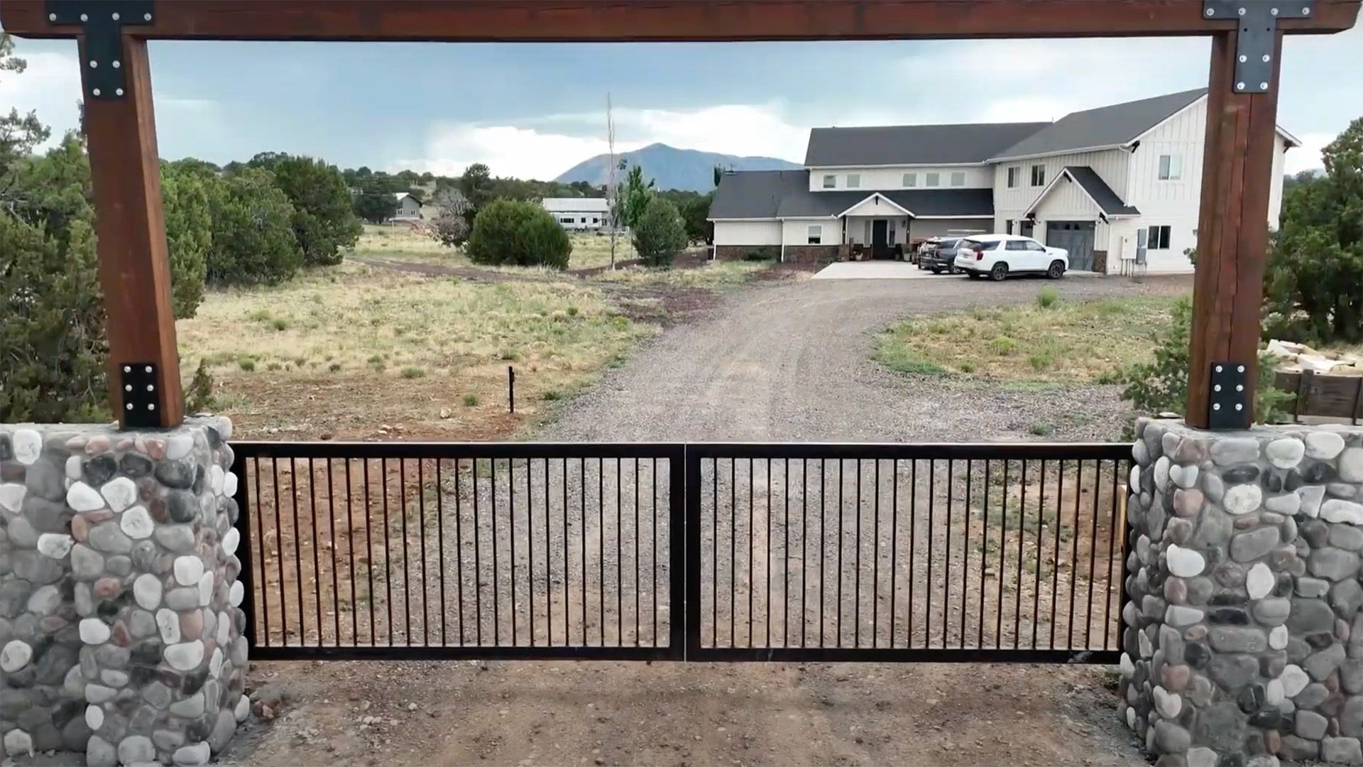 Wooden gate with stone pillars opening to a gravel drive and modern houses beyond