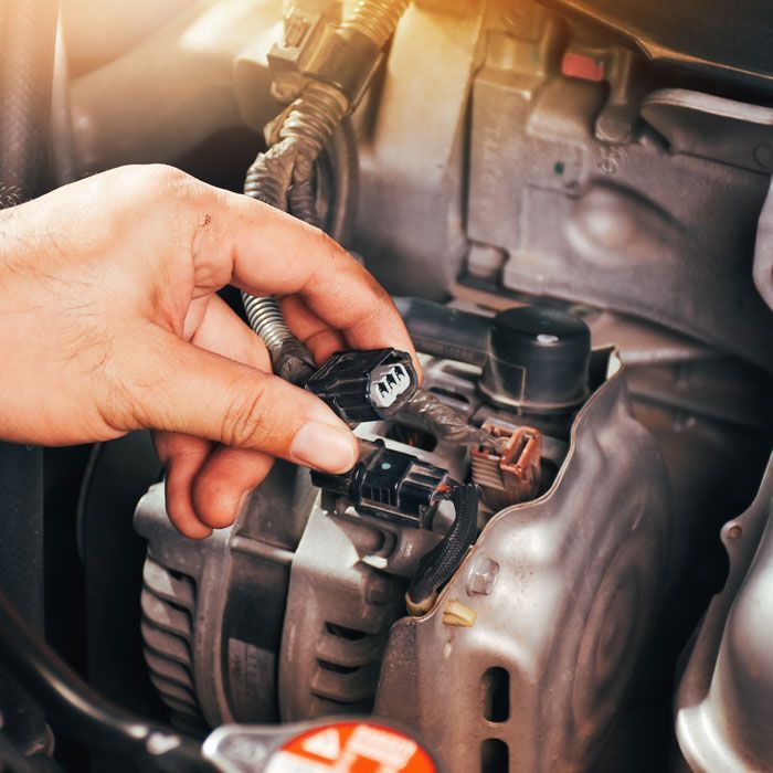 Hand holding a car engine connector near an alternator, with wiring and metal components visible.