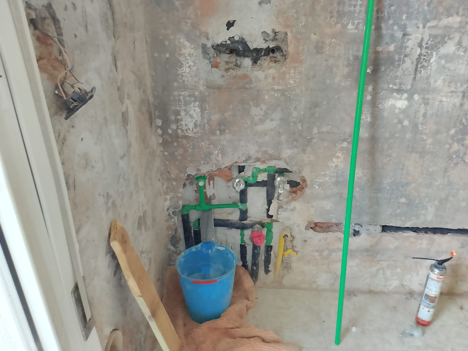 Parete interna danneggiata con tubi a vista, un secchio blu e una bomboletta spray sul pavimento.