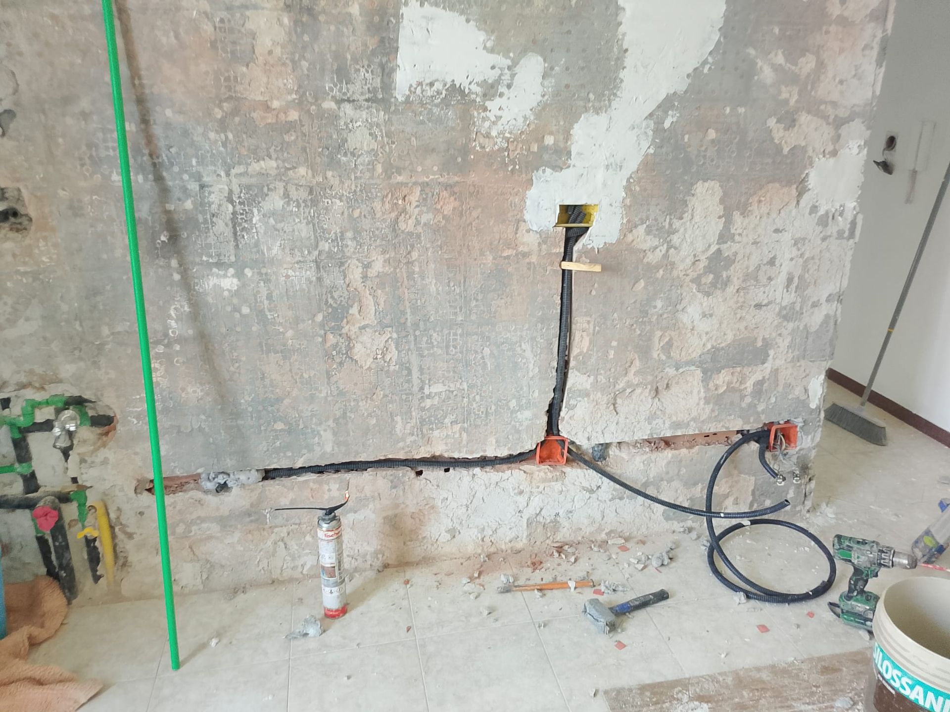 Cantiere edile con parete intonacata in modo grezzo, cavi elettrici a vista e attrezzi idraulici sul pavimento.