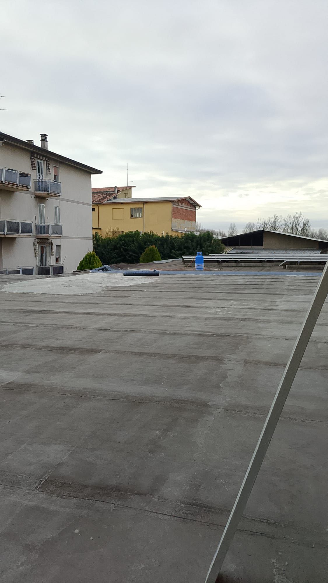 Tetto con superficie grigia e piatta, palazzi residenziali sulla sinistra e cielo nuvoloso sopra.