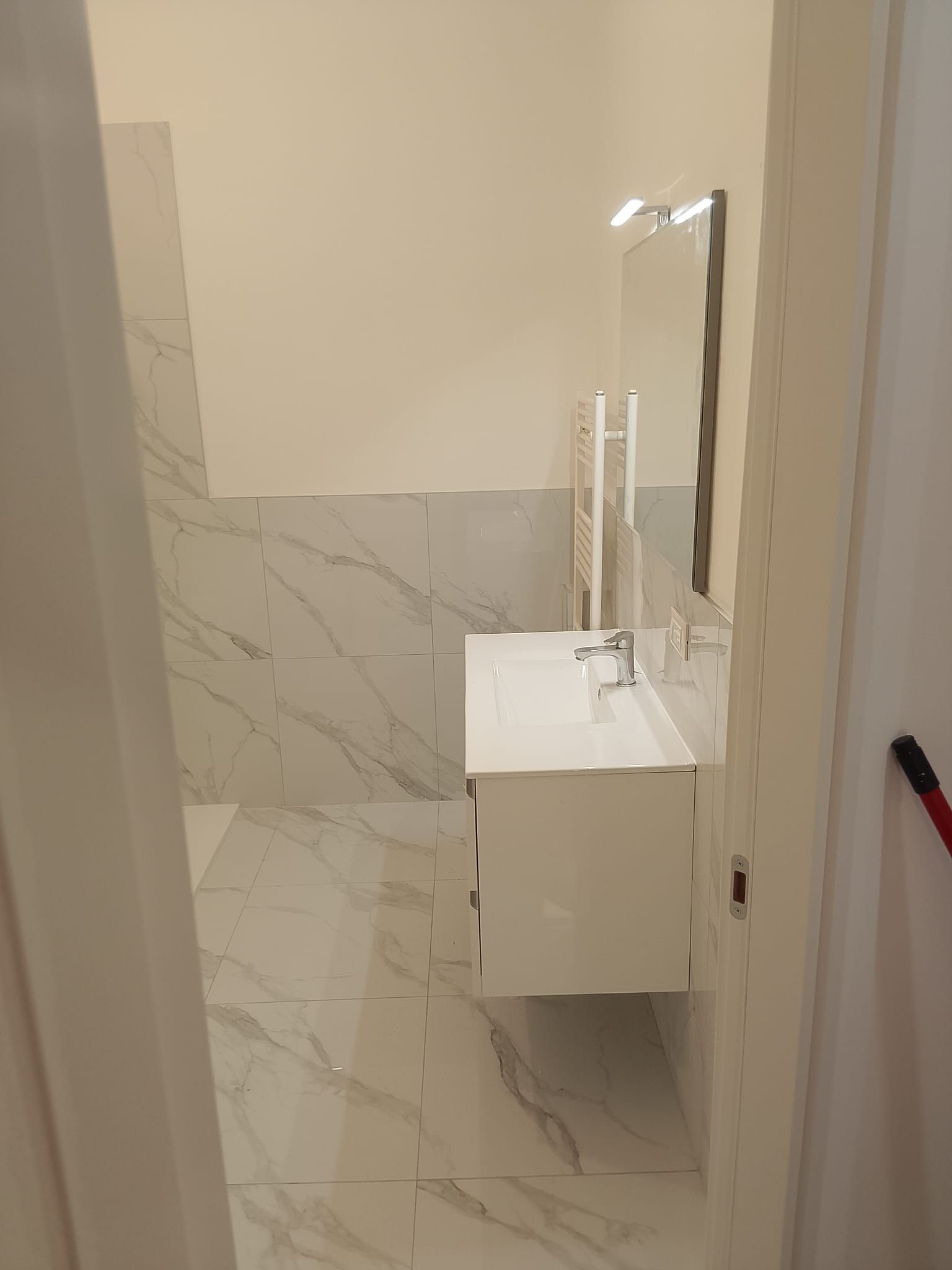Piccolo bagno con lavabo a parete, specchio e pareti e pavimento rivestiti con piastrelle bianche effetto marmo.