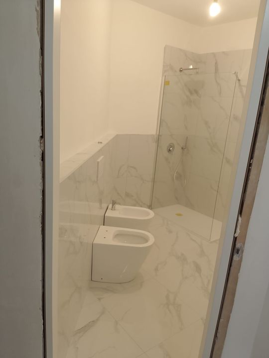 Piccolo bagno rivestito in marmo con bidet a parete, WC e box doccia in vetro.