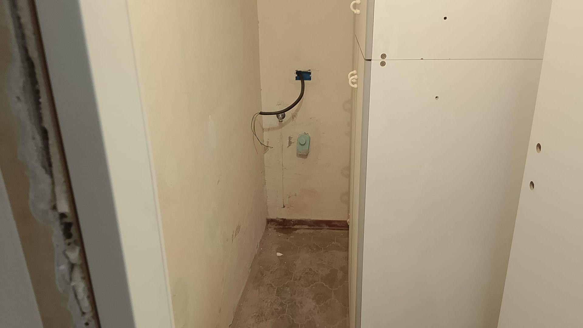 Stretto corridoio non rifinito con pareti spoglie, cavi elettrici a vista e una piccola presa a muro.