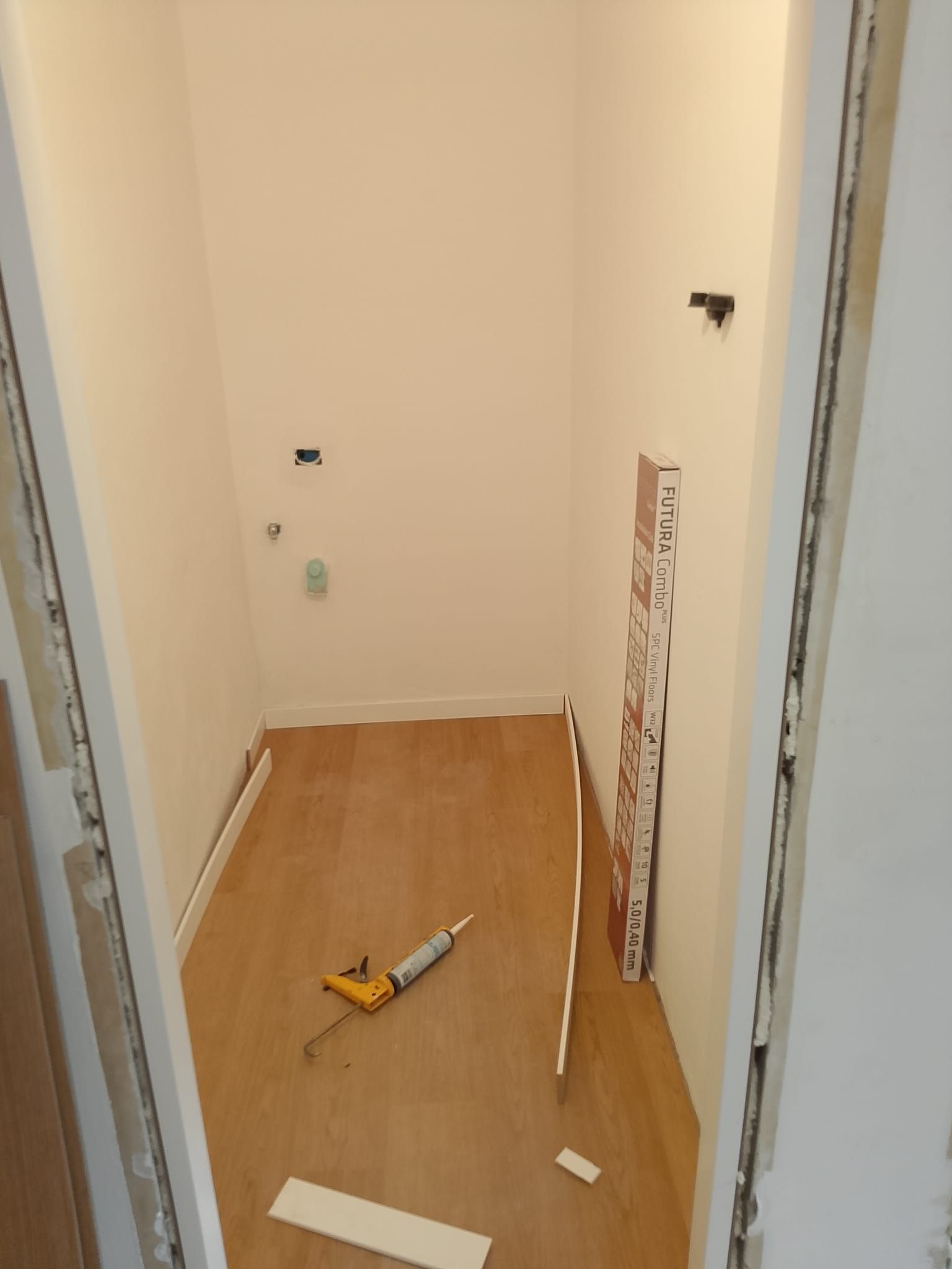 Corridoio stretto con attrezzi e detriti sul pavimento, pannello a muro non rifinito appoggiato alla porta.