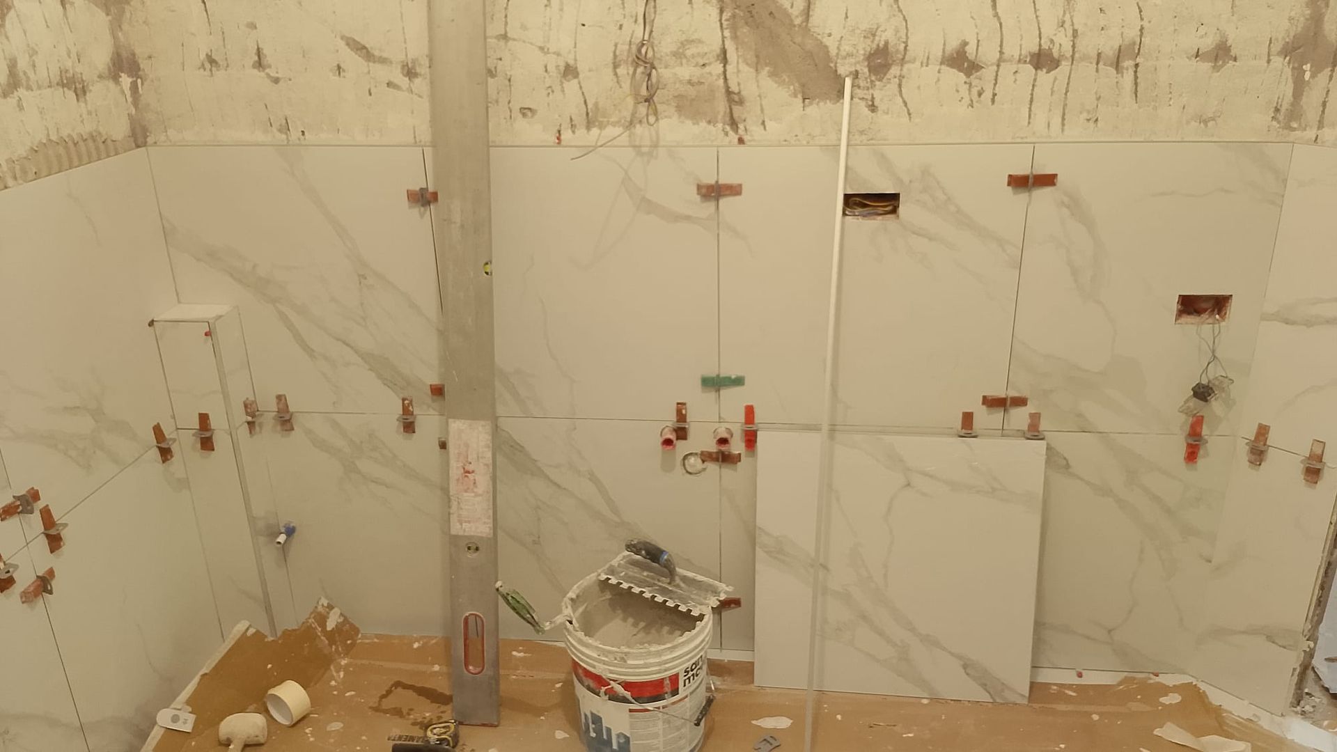Parete del bagno parzialmente piastrellata con piastrelle bianche effetto marmo, punti di colla a vista e un secchio sul pavimento.