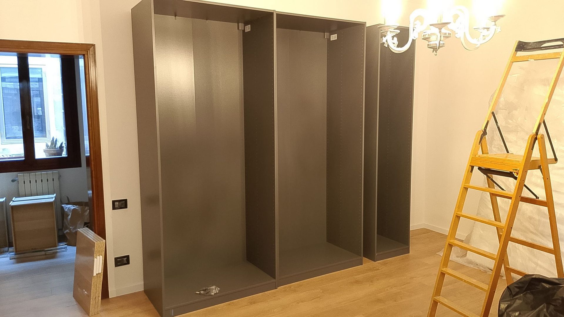 Armadi a muro grigi parzialmente assemblati in una stanza con pavimento in legno e una scala sulla destra.