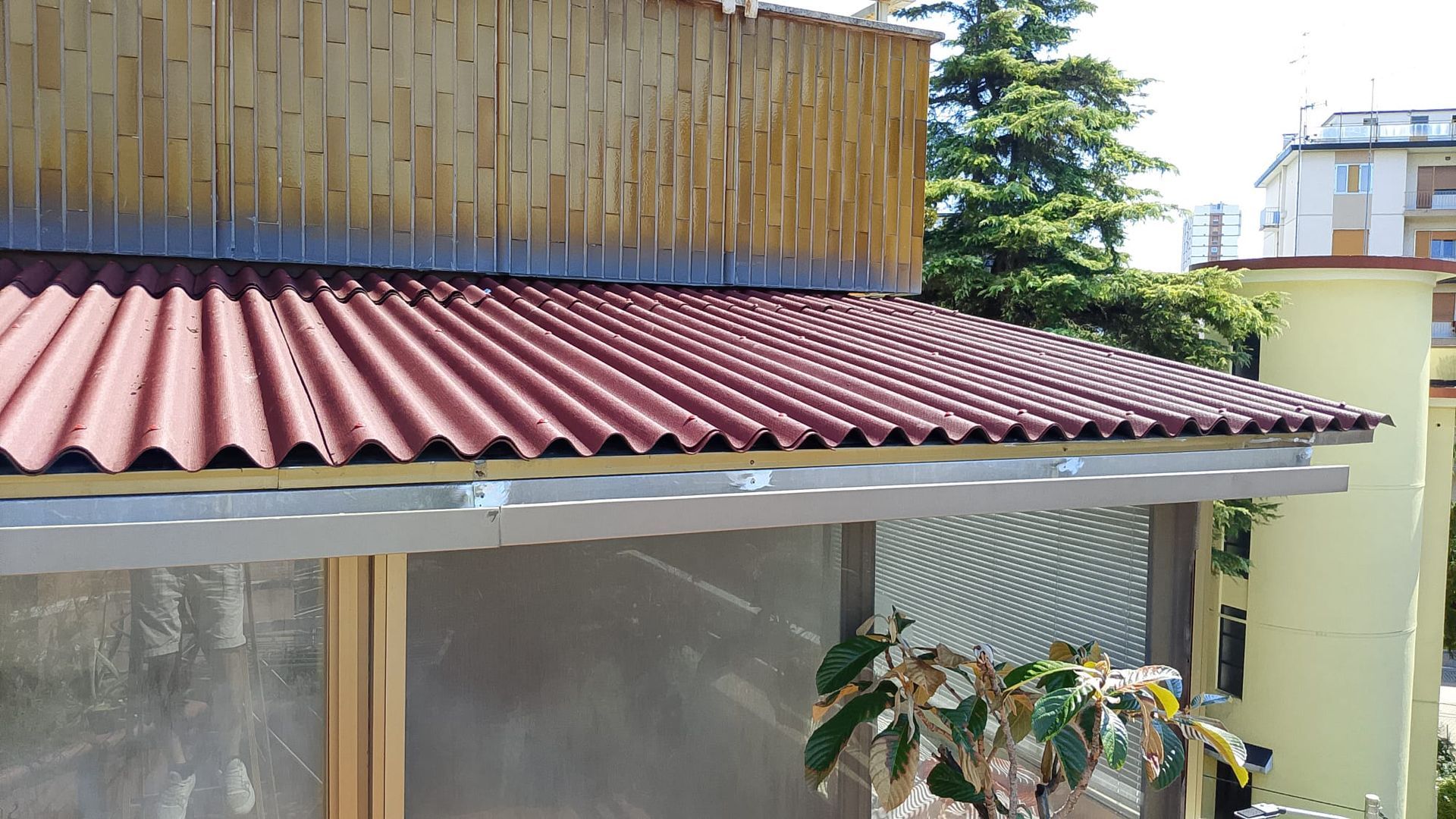 Tettoia con tenda da sole ondulata rossa, parete di bambù e pianta in vaso accanto a un edificio di colore chiaro.