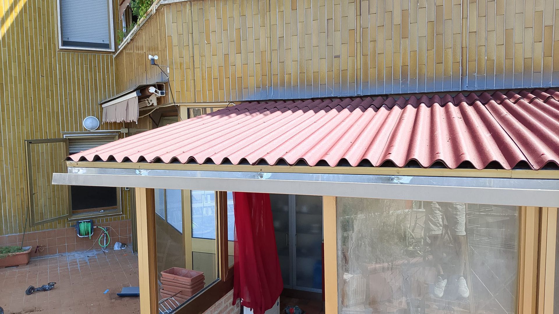 Piccolo patio in legno con tetto in lamiera ondulata rosa, tenda rossa e muro color beige dall'aspetto vissuto.