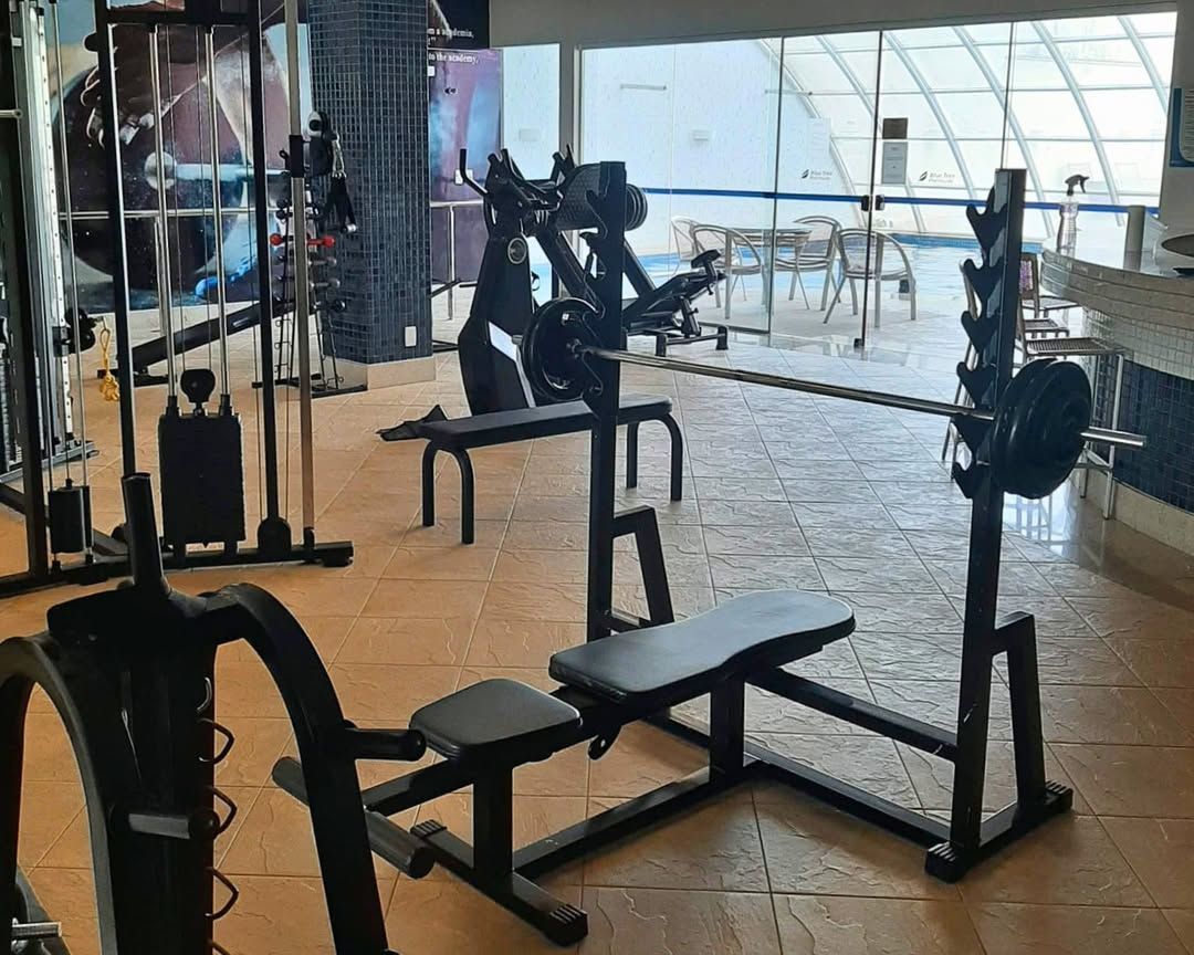 Academia com banco de musculação, suporte para agachamento e outros equipamentos de ginástica perto de uma grande janela com vista para um pátio.