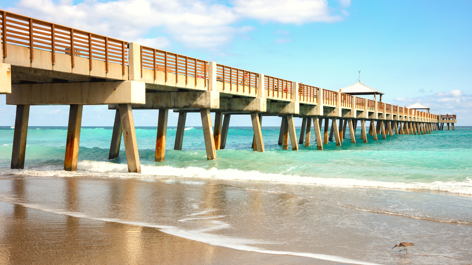 A long wooden pier extends over turquoise water on a sunny beach; clear blue sky.