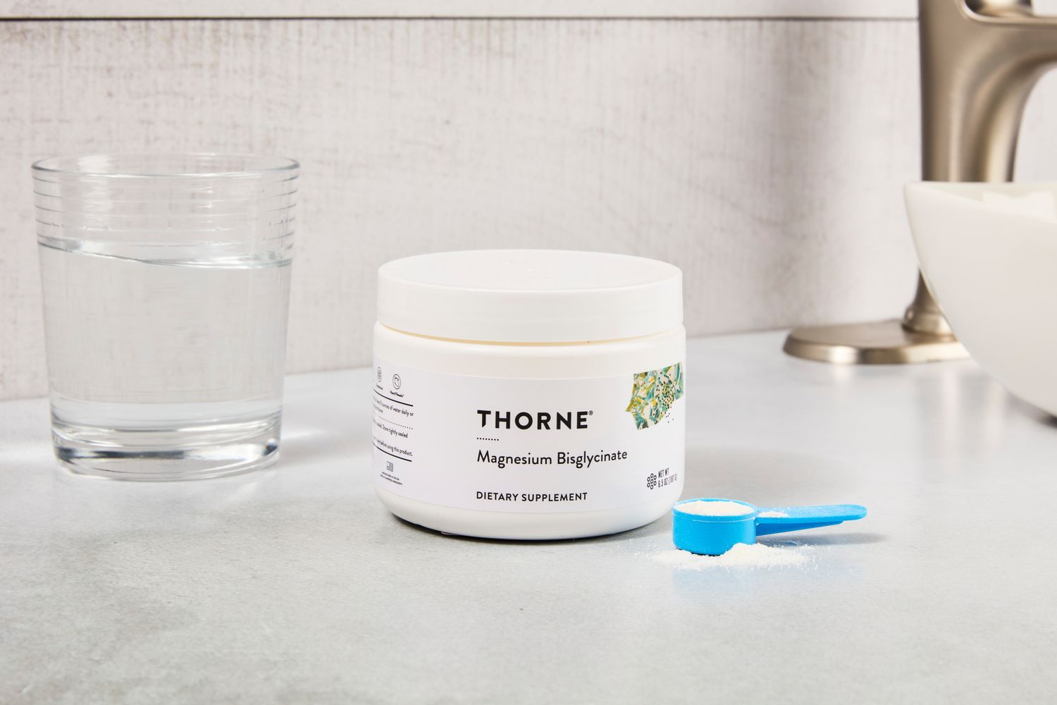 Jar of Thorne supplement next to a glass of water and a blue scoop on a counter.