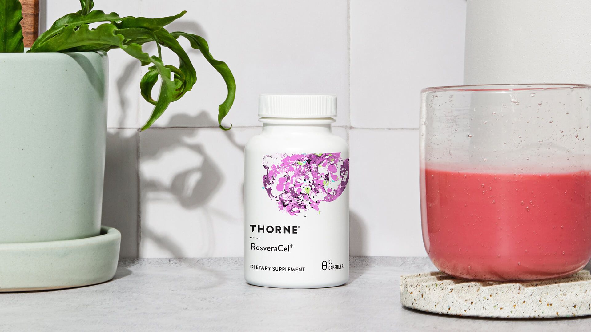 White bottle of Thorne supplements beside a pink drink in a glass; a plant in a light green pot is also visible.