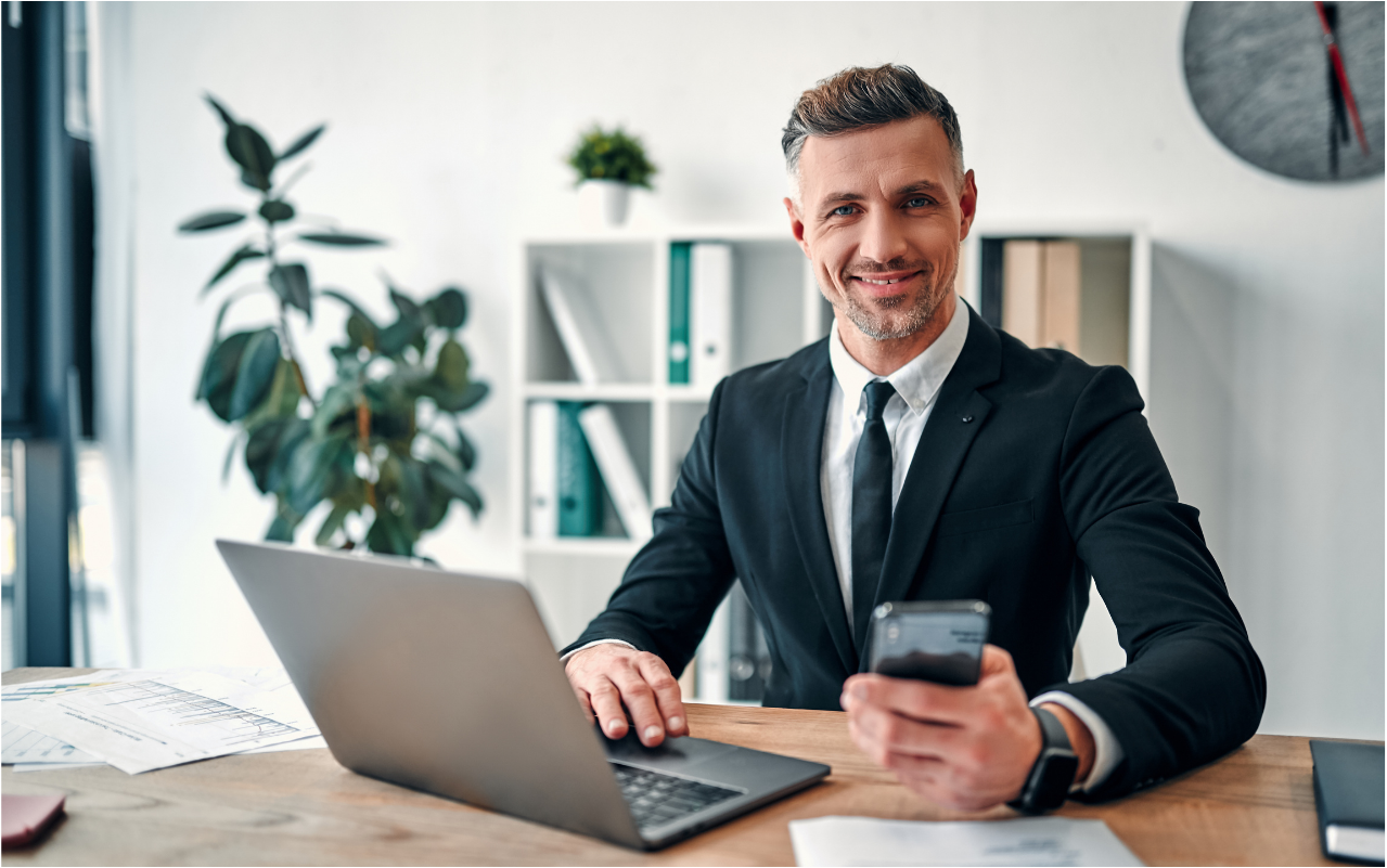 a man in a suit is sitting at a desk with a laptop and a cell phone.